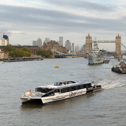 A modern ferry labeled "Uber Boat" travels on a river with Tower Bridge and city buildings in the background.