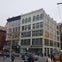 A multi-story building with large windows, located at a city intersection. People and vehicles are visible in the scene.