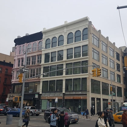A multi-story building with large windows, located at a city intersection. People and vehicles are visible in the scene.