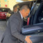 Two men in suits loading luggage into the trunk of a black SUV; other vehicles are parked in the background.