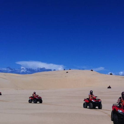 People riding red ATVs across a vast, sandy desert under a bright blue sky.
