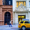 A street view showing a yellow taxi with advertisement, a pedestrian walking, and ornate building facades in the background.