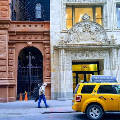 A street view showing a yellow taxi with advertisement, a pedestrian walking, and ornate building facades in the background.