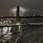 A nighttime view of a lit suspension bridge over a calm river, with reflections on the water and buildings in the background.