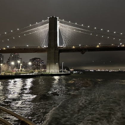 A nighttime view of a lit suspension bridge over a calm river, with reflections on the water and buildings in the background.