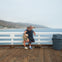 Two people stand on a wooden pier by the ocean, leaning on a white railing. One is taking a selfie. A trash bin is nearby.