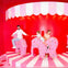 Three people, including a child, ride pink elephant-shaped carousel seats on a red and white striped stage with a scalloped canopy.