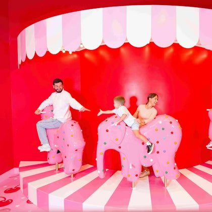 Three people, including a child, ride pink elephant-shaped carousel seats on a red and white striped stage with a scalloped canopy.