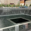A memorial fountain with water cascading into a square central void, surrounded by trees and urban buildings.