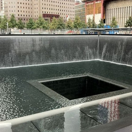 A memorial fountain with water cascading into a square central void, surrounded by trees and urban buildings.