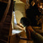 A woman leans into an art installation, inspecting illuminated smoke under a spotlight, surrounded by intricate wooden structures.