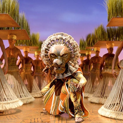 A performer in a lion costume leads a group of dancers in an elaborate stage setting with tall grass props.