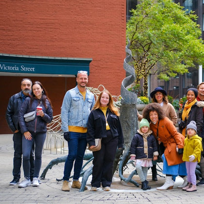 A group of people posing together outdoors in front of a sculpture, with a brick building and trees visible in the background.