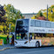 Double-decker tour bus parked near a park with lush trees and a building in the background. Several people are nearby.