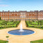 A grand red brick building with numerous windows overlooks a formal garden featuring a circular fountain and manicured lawns.