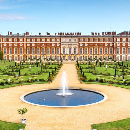A grand red brick building with numerous windows overlooks a formal garden featuring a circular fountain and manicured lawns.