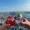 Two hands holding drinks with "Edge New York" glasses, overlooking a cityscape and river under a clear sky.