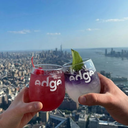 Two hands holding drinks with "Edge New York" glasses, overlooking a cityscape and river under a clear sky.