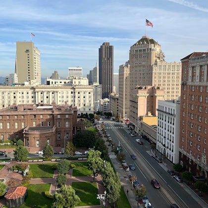 Cityscape showing a park, buildings, and a street with cars under a blue sky.