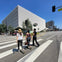 Three people crossing a street at a crosswalk, with a modern white building and traffic light visible in the background.