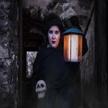 Person dressed in dark clothing holds a lit lantern and a skull inside an old, stone structure with a mountainous background.