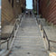 Two people climbing long concrete stairs with handrails on both sides, flanked by brick and wooden walls, under a clear sky.