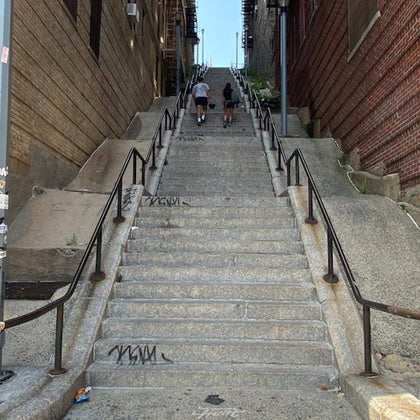 Two people climbing long concrete stairs with handrails on both sides, flanked by brick and wooden walls, under a clear sky.