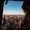 A person in a helicopter overlooking a city skyline during sunset with buildings and a clear sky visible.