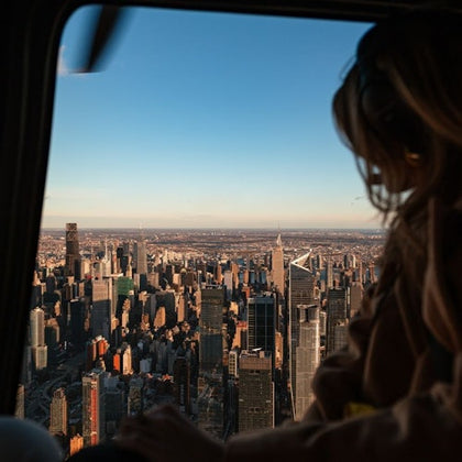 A person in a helicopter overlooking a city skyline during sunset with buildings and a clear sky visible.