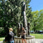 A person speaks to two individuals in front of a totem pole in a park setting with trees and a clear sky in the background.