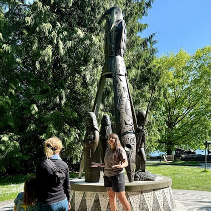 A person speaks to two individuals in front of a totem pole in a park setting with trees and a clear sky in the background.