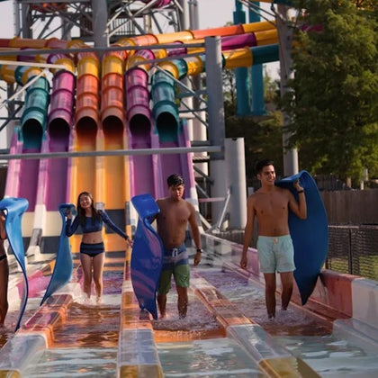 Four people holding mats walk through shallow water at the end of colorful water slides in an outdoor water park.