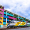 A modern Brightline train passes by a multicolored building with murals and palm trees under a cloudy sky.