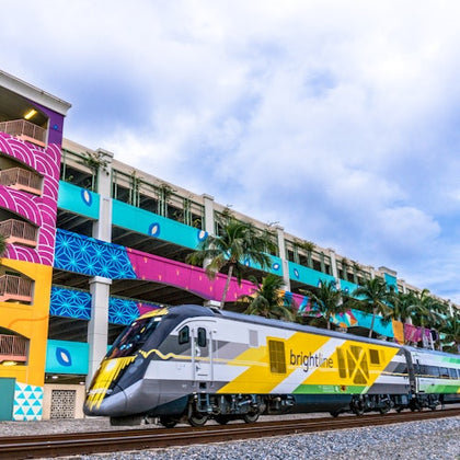 A modern Brightline train passes by a multicolored building with murals and palm trees under a cloudy sky.