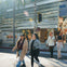 Four women walking across a sunny street in front of a Louis Vuitton store, with palm trees and reflections in the glass.