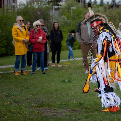 A person in traditional regalia performs a dance outdoors while several onlookers in casual attire watch.