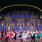 A group of performers in elegant attire dancing on a theater stage with an ornate backdrop and chandeliers.