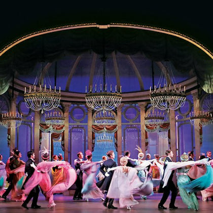 A group of performers in elegant attire dancing on a theater stage with an ornate backdrop and chandeliers.
