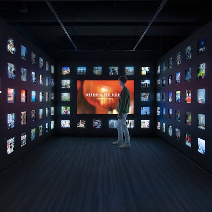 A person stands in a dimly lit exhibit room with illuminated screens displaying various images on the walls.