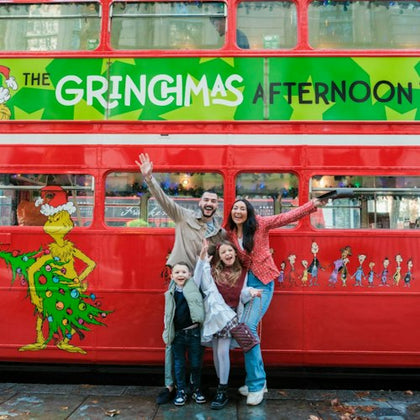 A family of four posing happily in front of a red bus decorated with "The Grinchmas Afternoon" and Grinch-themed artwork.