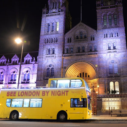 A yellow double-decker bus with "See London by Night" text, parked in front of an illuminated historic building at night.