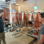 Two people in a brewery standing near large copper brewing tanks and equipment, surrounded by hoses and pipes.