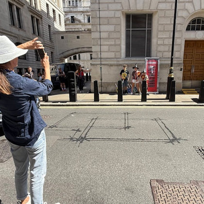 A person in a hat taking a photo of a group posing near a red phone booth on a sunny city street.