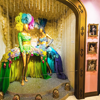 Two mannequins dressed in colorful, feathered showgirl costumes displayed in an ornate window with framed photos on the side.