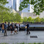 A group of people walking near a fountain surrounded by trees and modern buildings in a city park.