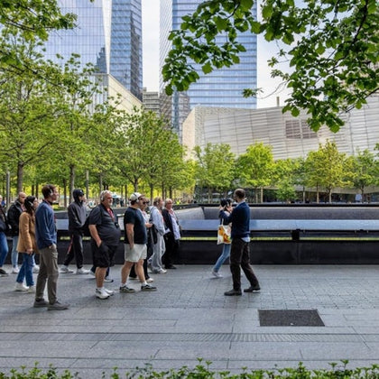 A group of people walking near a fountain surrounded by trees and modern buildings in a city park.