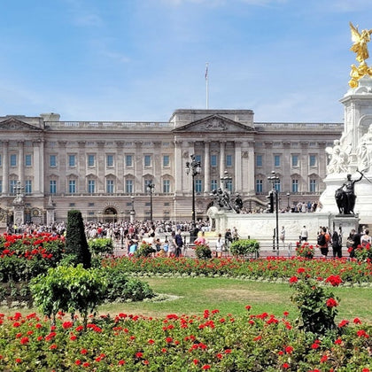 Buckingham Palace with crowds of people and the Victoria Memorial in front, surrounded by vibrant red flowers and greenery.