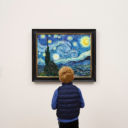 A child in a blue vest observes Van Gogh's 'Starry Night' painting displayed on a white wall in a gallery.