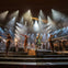 A theatrical performance on a stage with actors singing and dancing under dramatic lighting.