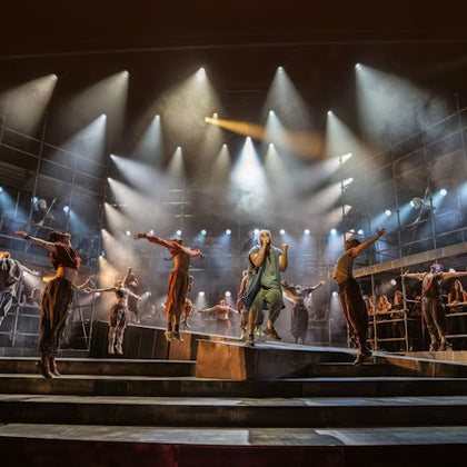 A theatrical performance on a stage with actors singing and dancing under dramatic lighting.